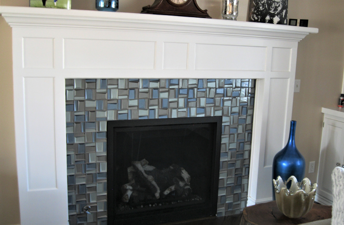 A white wooden mantelpiece above a fireplace with a mosaic tile surround in shades of blue and gray. Decorative vases are placed on the mantel and to the right of the fireplace.